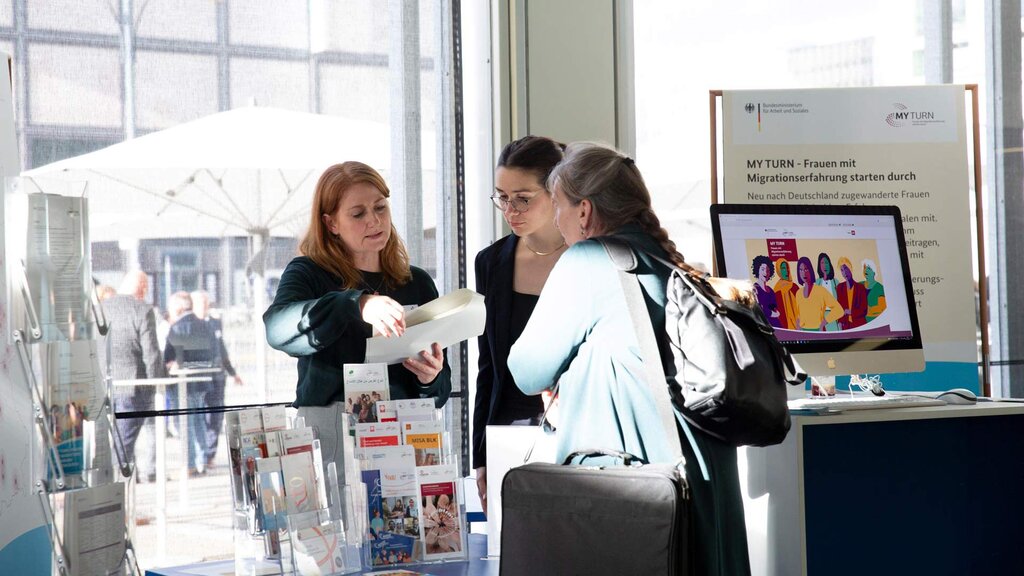 Eine Mitarbeiterin des MY TURN-Projektes berät zwei Informationssuchende auf dem Tag der Jobcenter